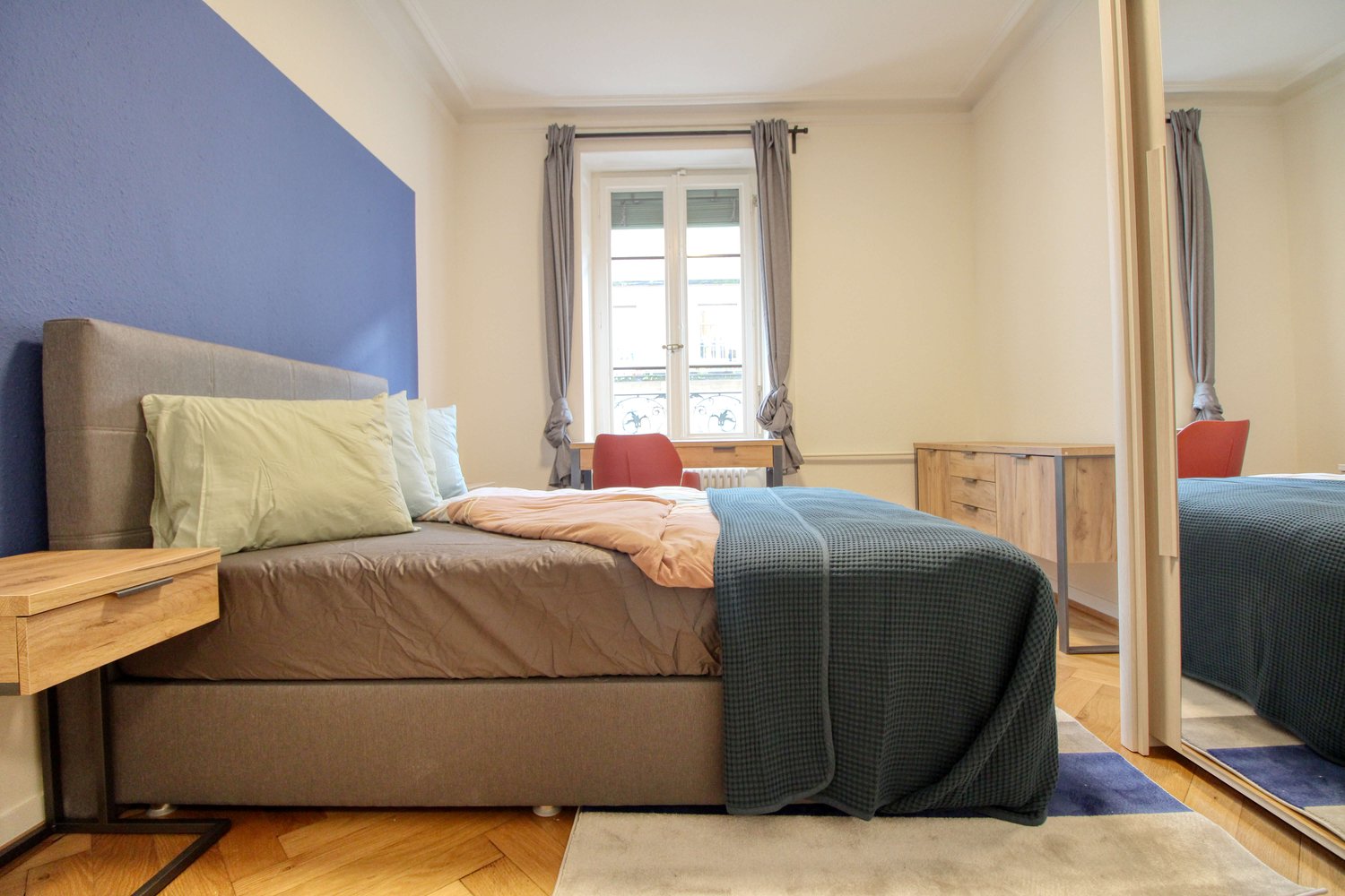 double bed, wooden nightstand, blue wall, window with balcony, wooden wardrobe, radiator, desk with chair, mirror