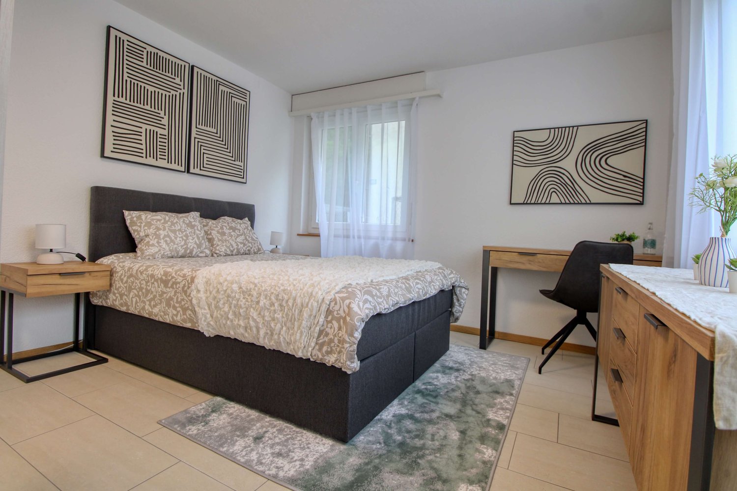 spacious bedroom with a double bed, bedside tables, lamp, desk, black chair, flower vase, wooden cabinet, artwork on the wall, rug, and large window with curtains