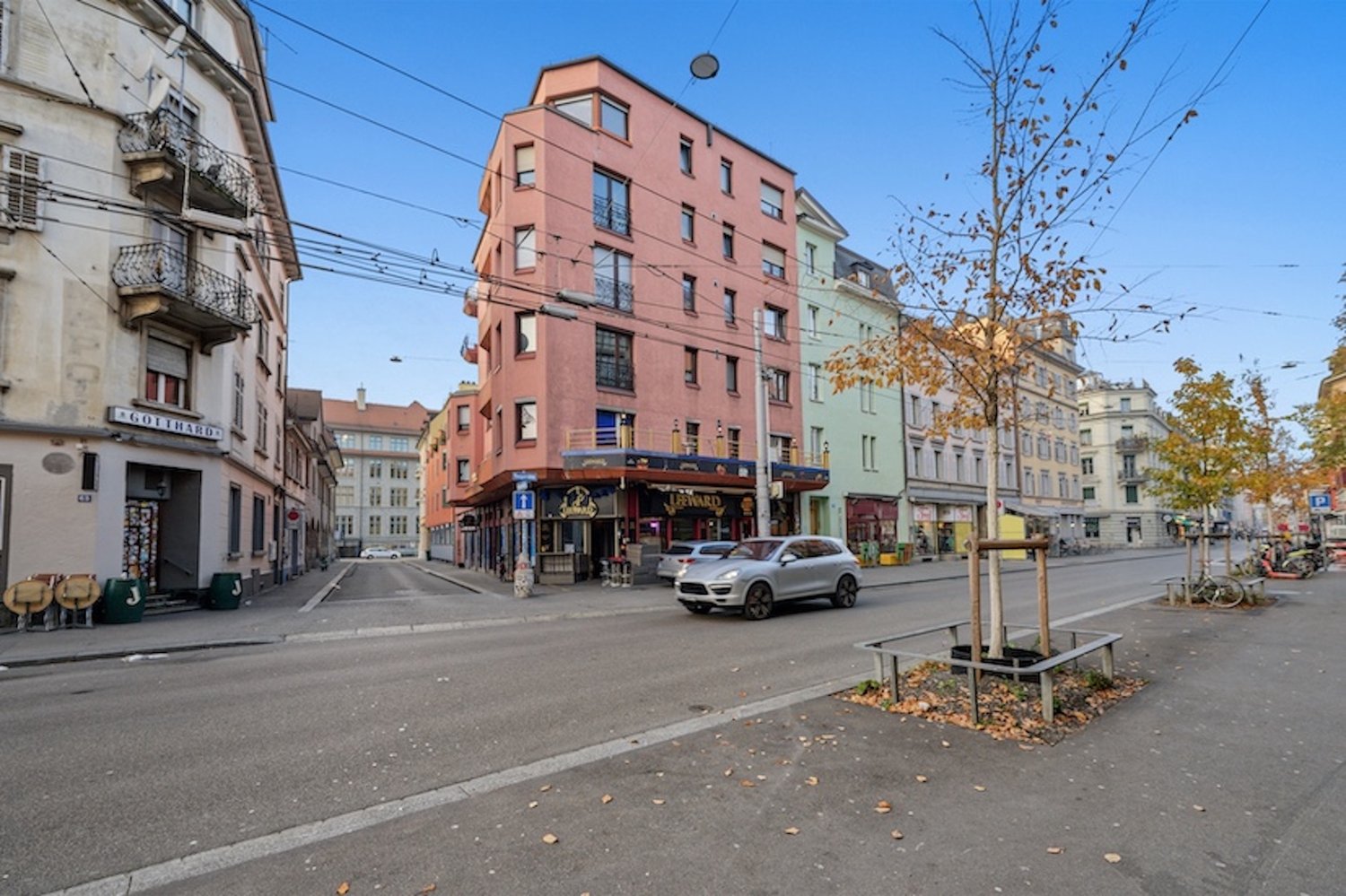 City street with a pink building, gray car, bicycles, trees, parked cars, sign on building.