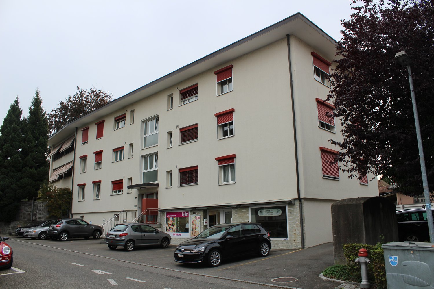 An apartment building with red shutters, several cars parked in front, ground floor business space