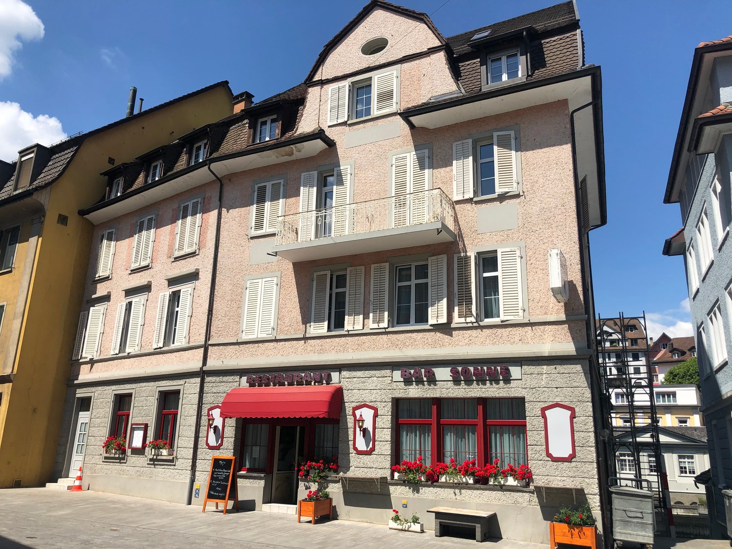 Building facade with white shutters, red awning, windows with flower boxes, entrance with sign, balcony, and sign