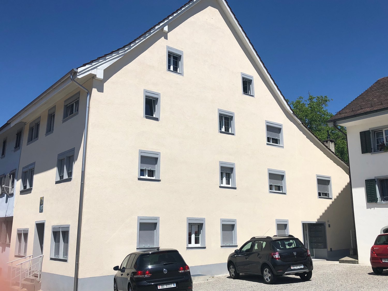A multi-story residential building with a light-colored exterior, multiple windows, and parked cars in the foreground.