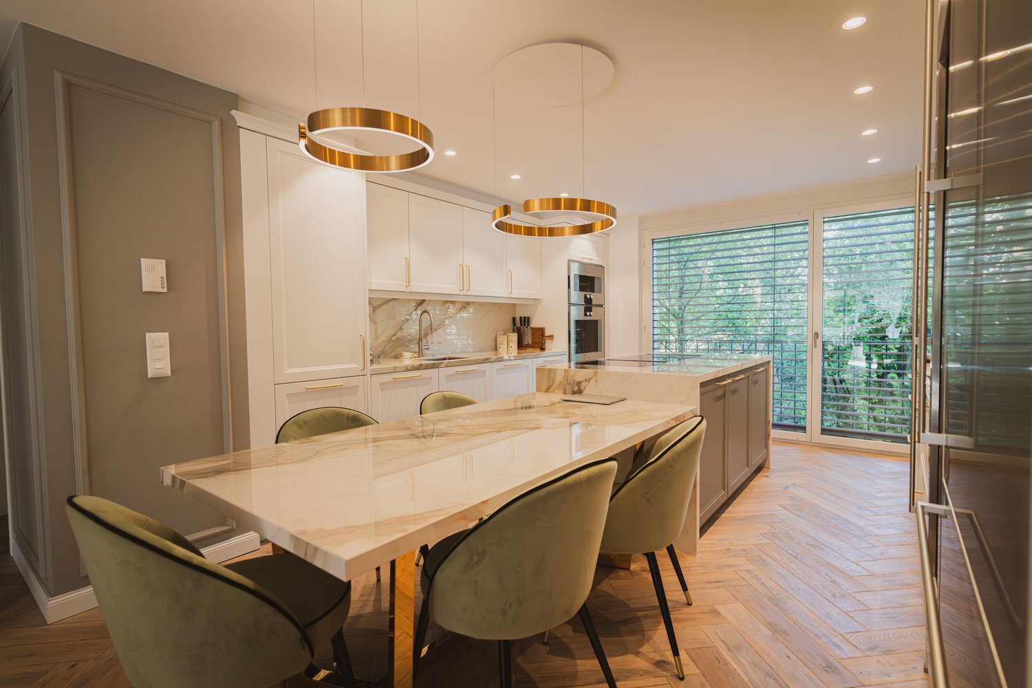 Spacious open-concept kitchen and dining area with modern, high-end finishes. Large marble-topped dining table with plush green chairs, two gold-rimmed pendant lights above. Fully equipped kitchen with white cabinets, stainless steel appliances, and a vie