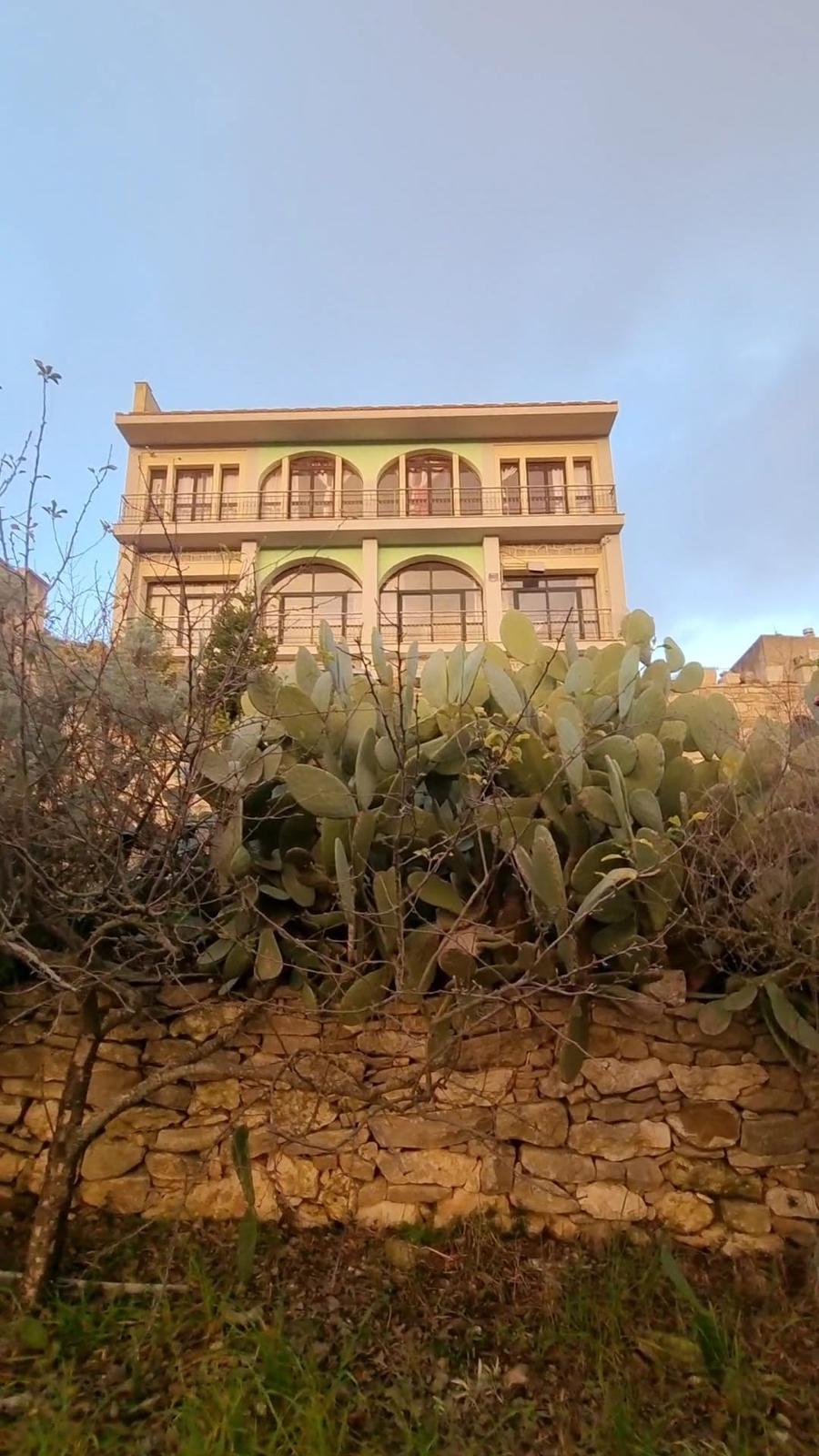 Two-story building with arched windows and balconies, surrounded by a stone wall and cactus plants in the foreground.