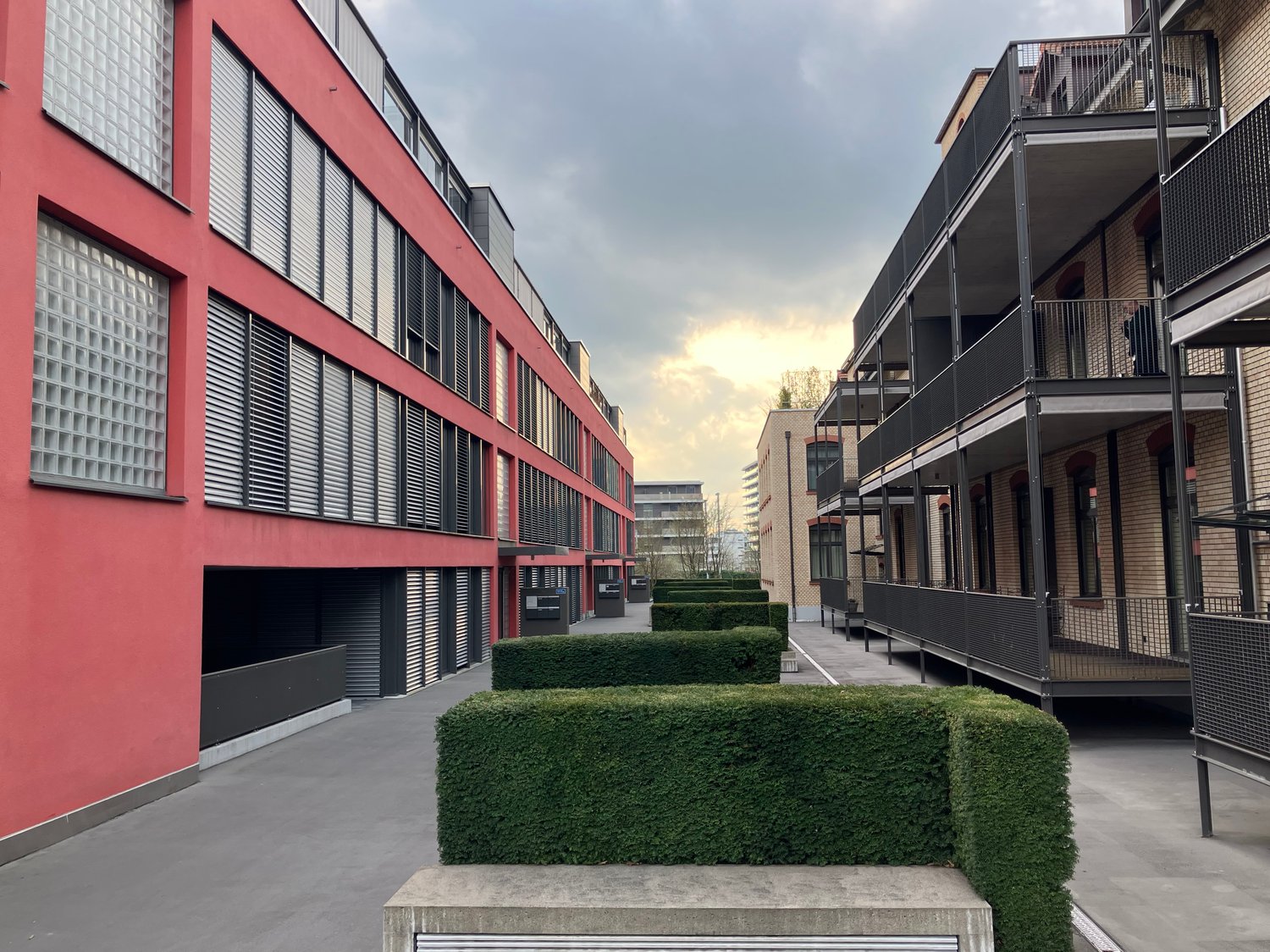 Modern red buildings with large windows, two balconies on the right side, concrete walkway, hedges, and grey concrete bases.