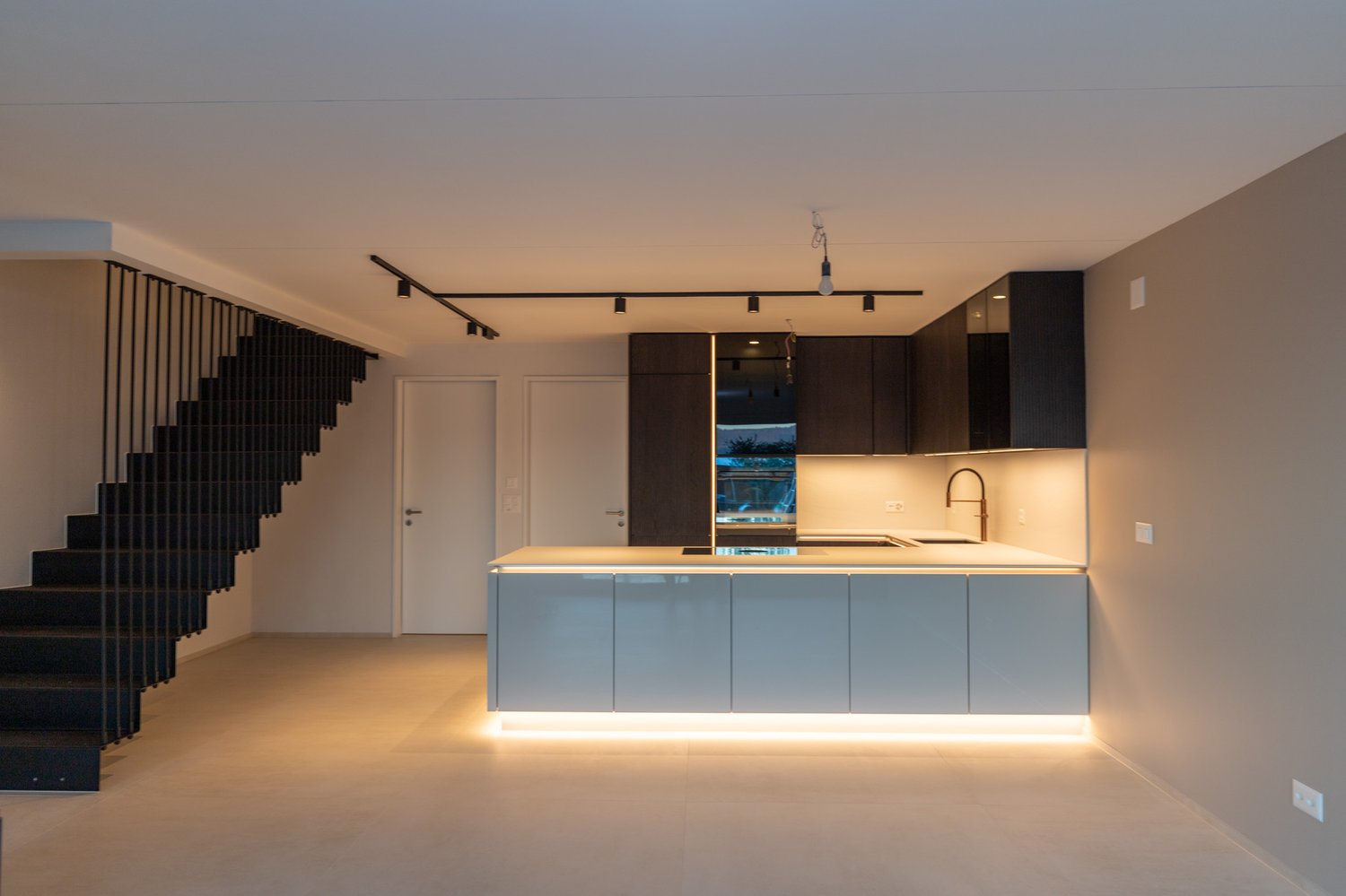 Modern kitchen with open floor plan, sleek black metal staircase, white countertops, integrated sink, and recessed lighting.