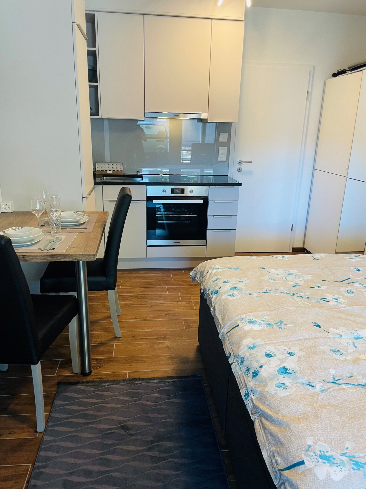The image shows a studio apartment with an open-plan layout. The kitchen area features white cabinets, a built-in oven, and a dishwasher. There is a dining table with chairs near the kitchen. The living area has a bed with a floral patterned bedding. The