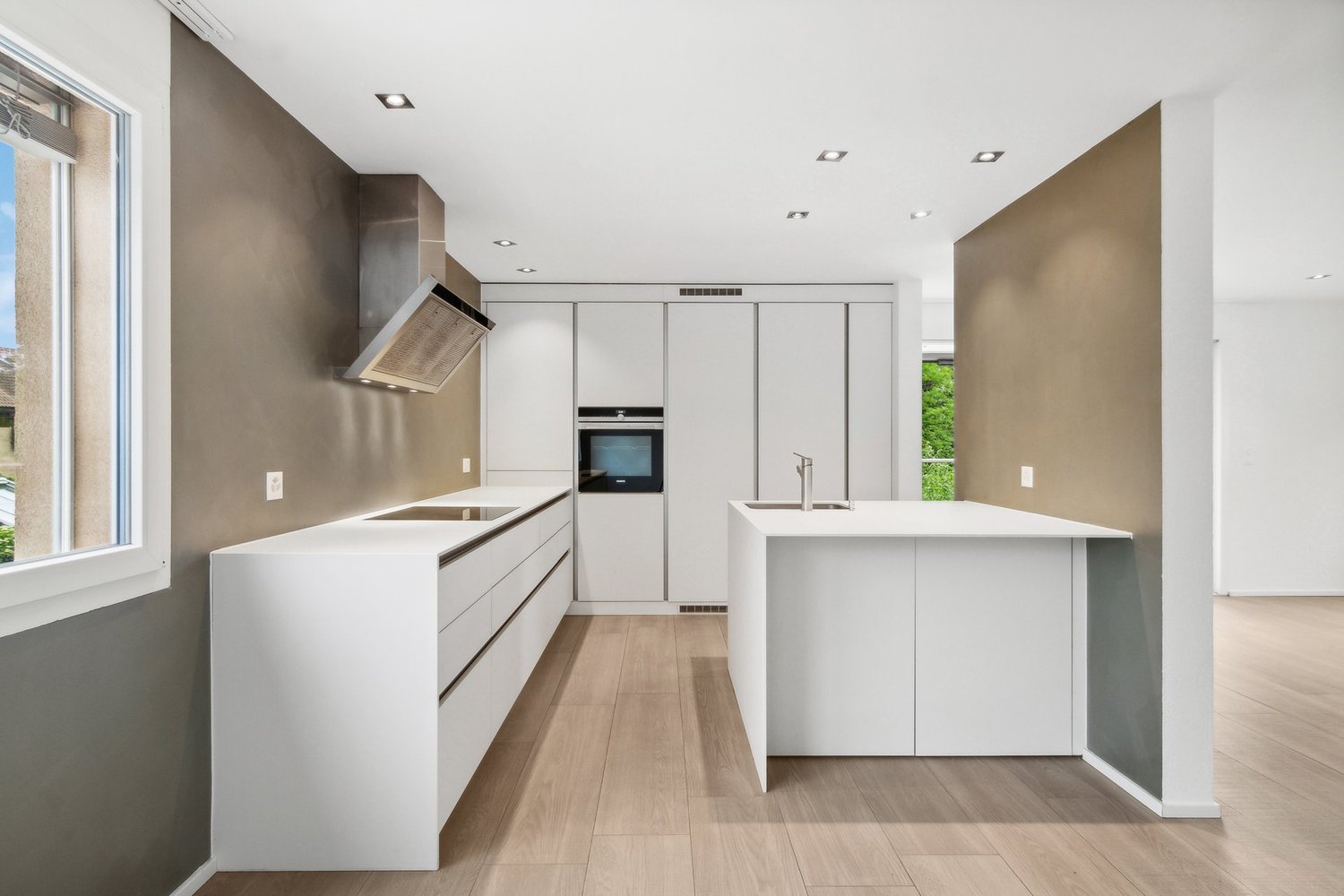 Modern, minimalist kitchen with white cabinets, built-in appliances, and hardwood flooring. The kitchen features a central island with a sink and countertop space. The walls are painted in a neutral, earthy tone, and the lighting is recessed.