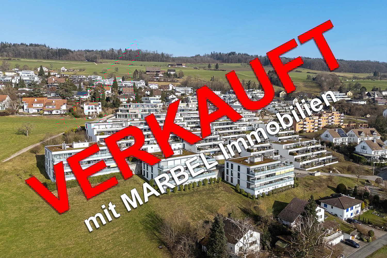 The image shows an aerial view of a town with a mix of residential and commercial buildings. The town is situated in a hilly, forested area with a clear blue sky in the background. The buildings appear to be a mix of modern and traditional architectural s