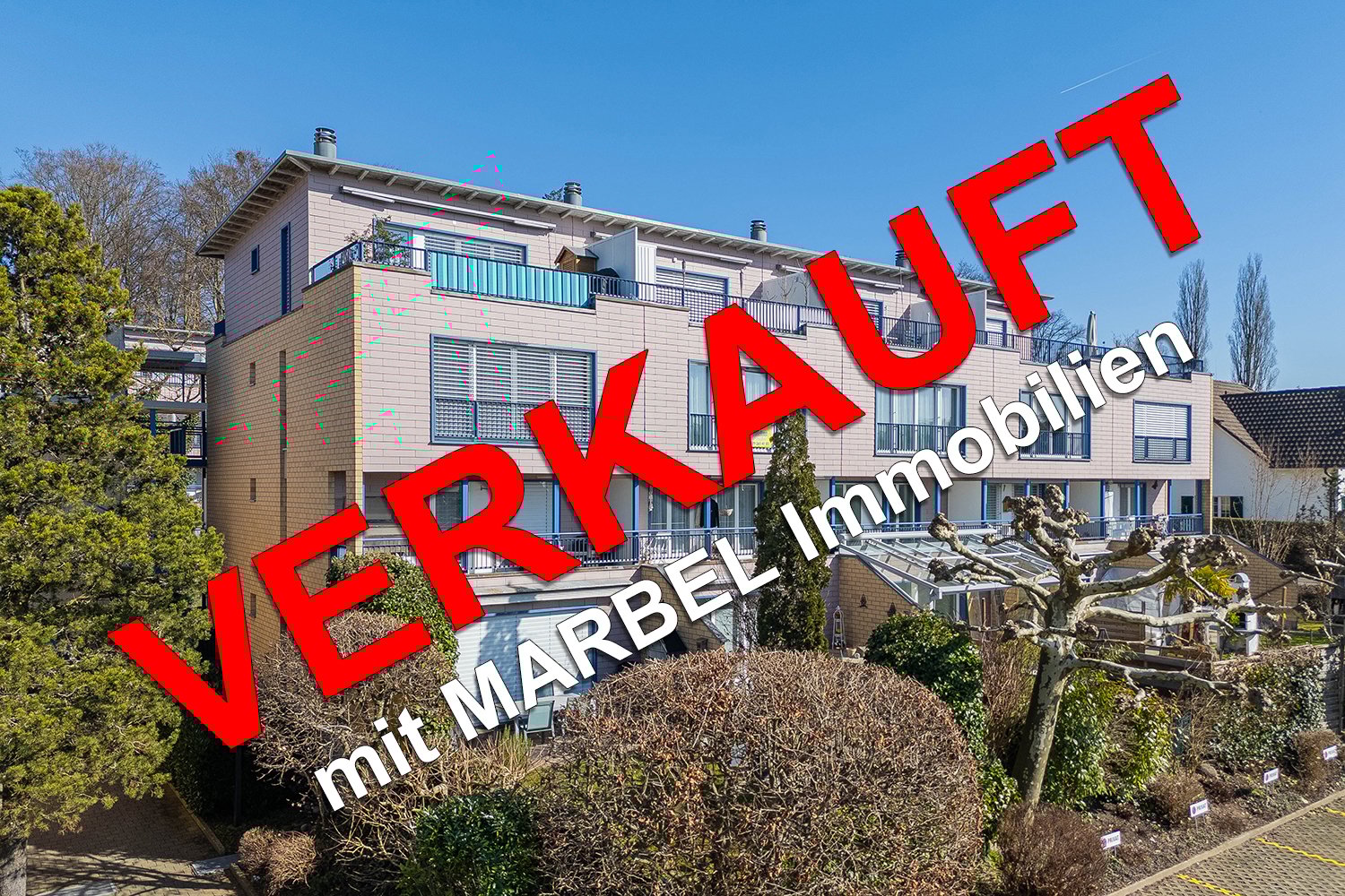 A multi-story apartment building with balconies, located in a residential area with trees and greenery surrounding it.