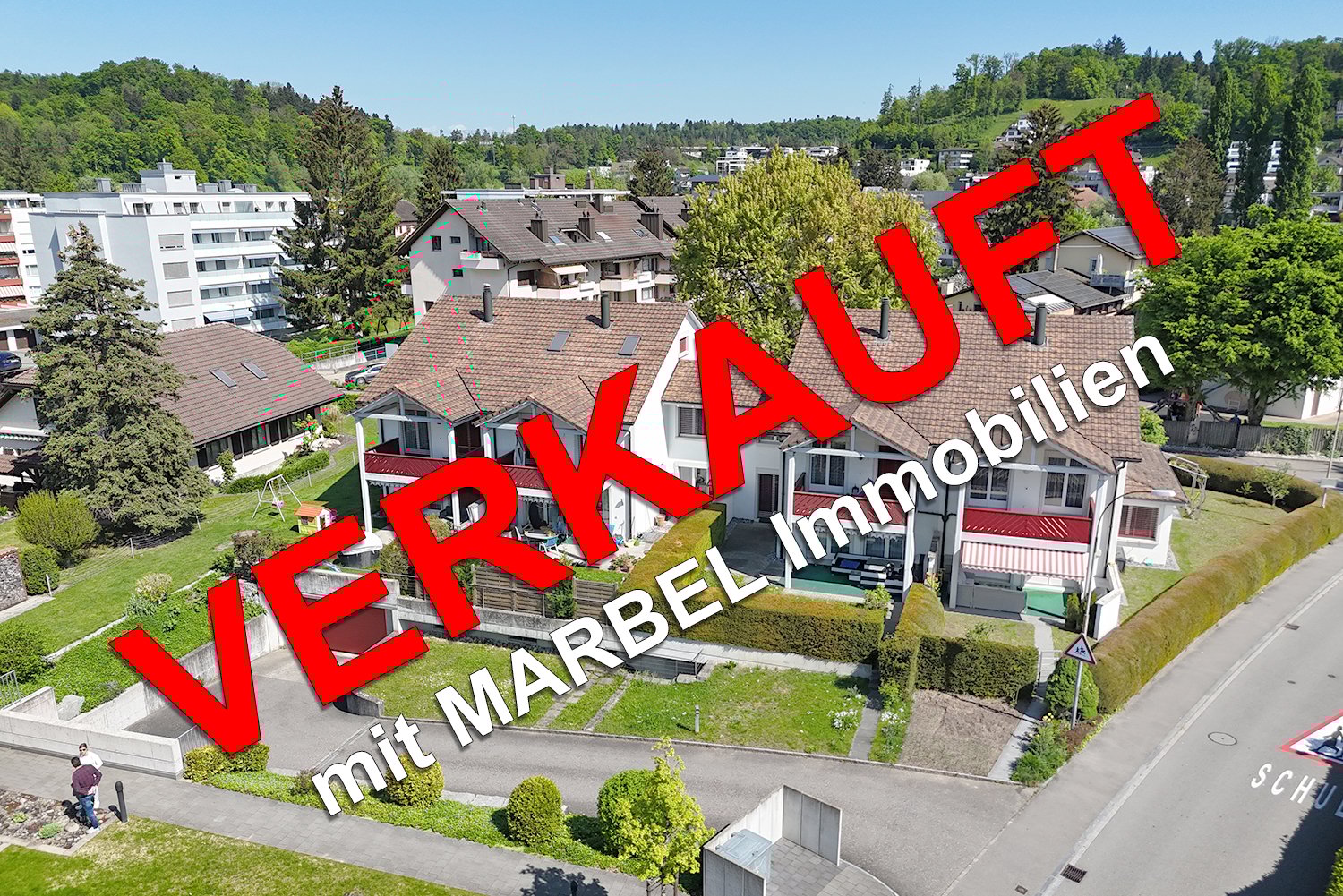 The image shows a residential area with several multi-story apartment buildings surrounded by trees and greenery. The buildings have red tile roofs and there appears to be a parking area visible in the foreground.