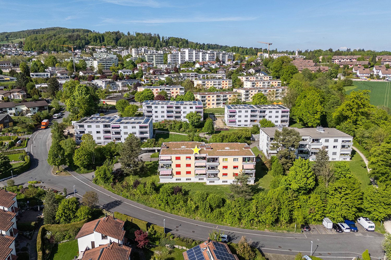 residential buildings, solar panels on roofs, road intersection, cars parked, greenery and trees, mountainous background, large area of green field, yellow star on a building
