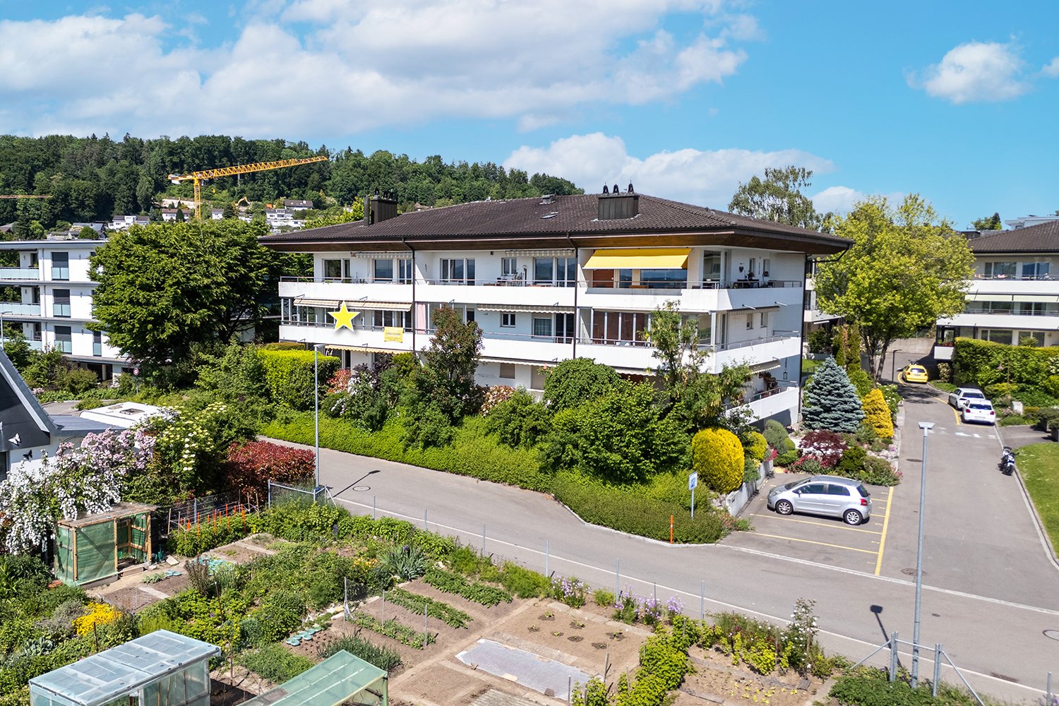 White building with yellow awnings, parked cars, road, garden, trees, buildings in the background