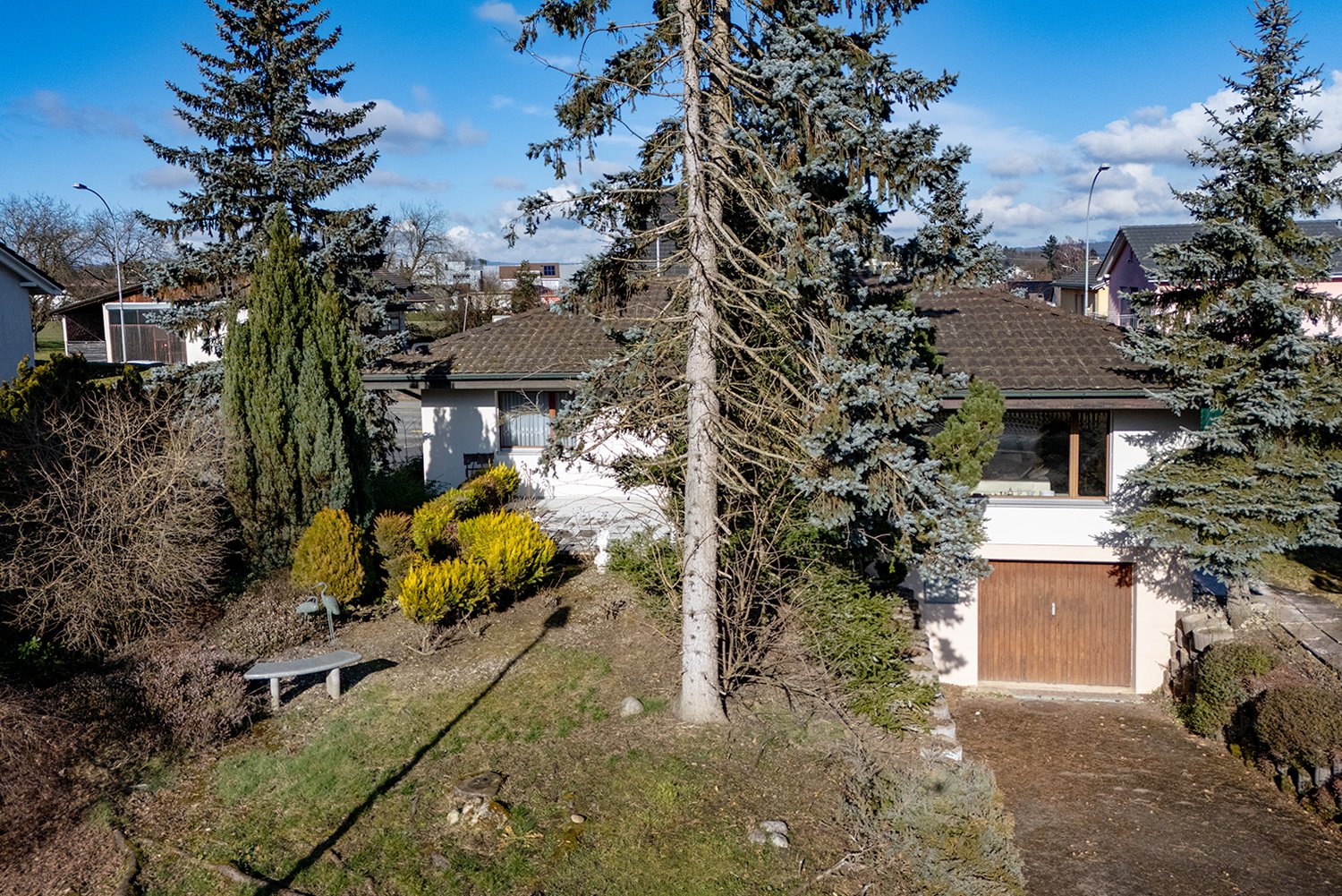 Detached house, gable roof, white walls, brown roof, wooden garage, trees, bushes, bench