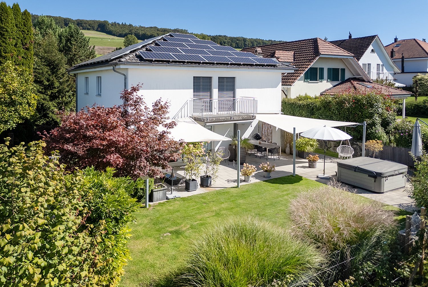 A modern white house with a roof-mounted solar panel system, a spacious backyard with outdoor seating and a jacuzzi, surrounded by greenery.