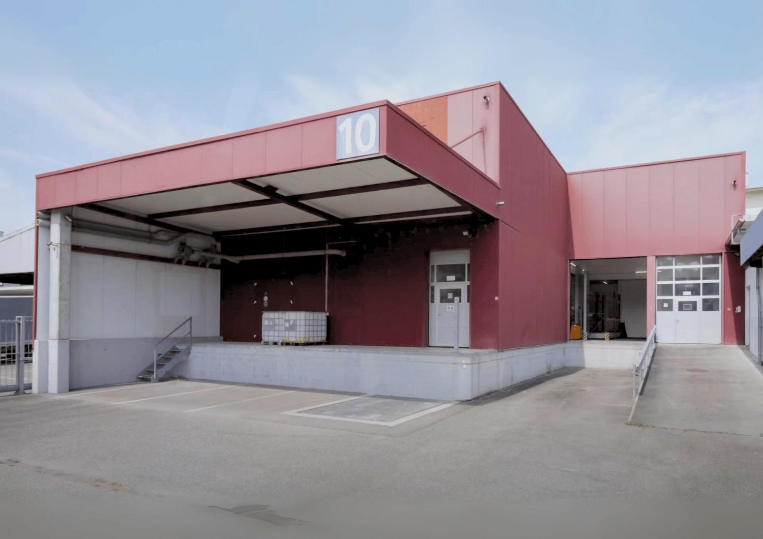 Warehouse: large open entrance, red walls, white ceiling, two entry doors, storage container, fenced parking lot
