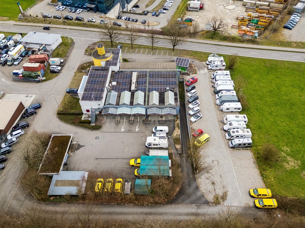 Aerial view of a parking lot with multiple vehicles parked, a building with solar panels, and a road with cars.