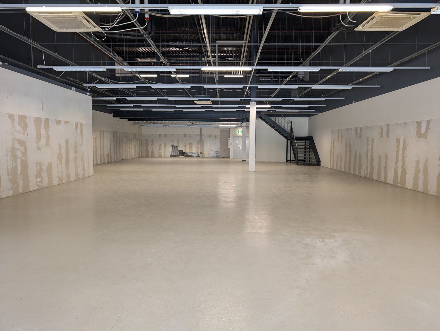 A vacant commercial space with gray concrete flooring, white walls, exposed beams, and overhead fluorescent lighting. There is a staircase in the right corner and an entrance at the back.
