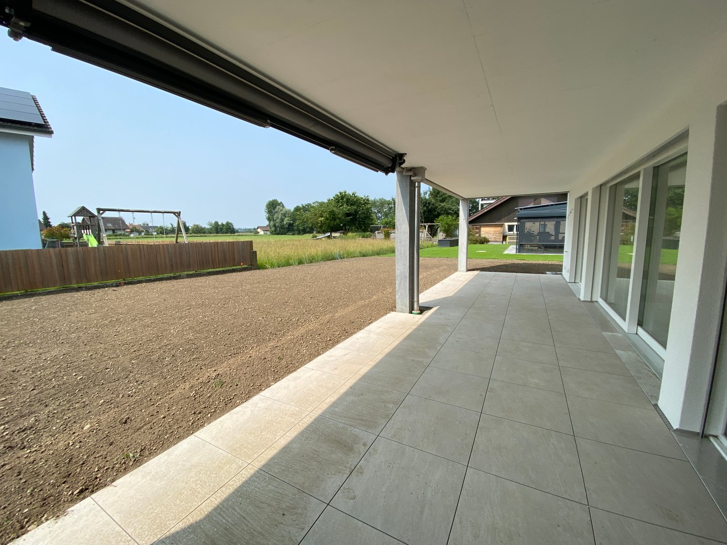 An outdoor balcony with tile flooring, large windows, and a view of a grassy field and a fence