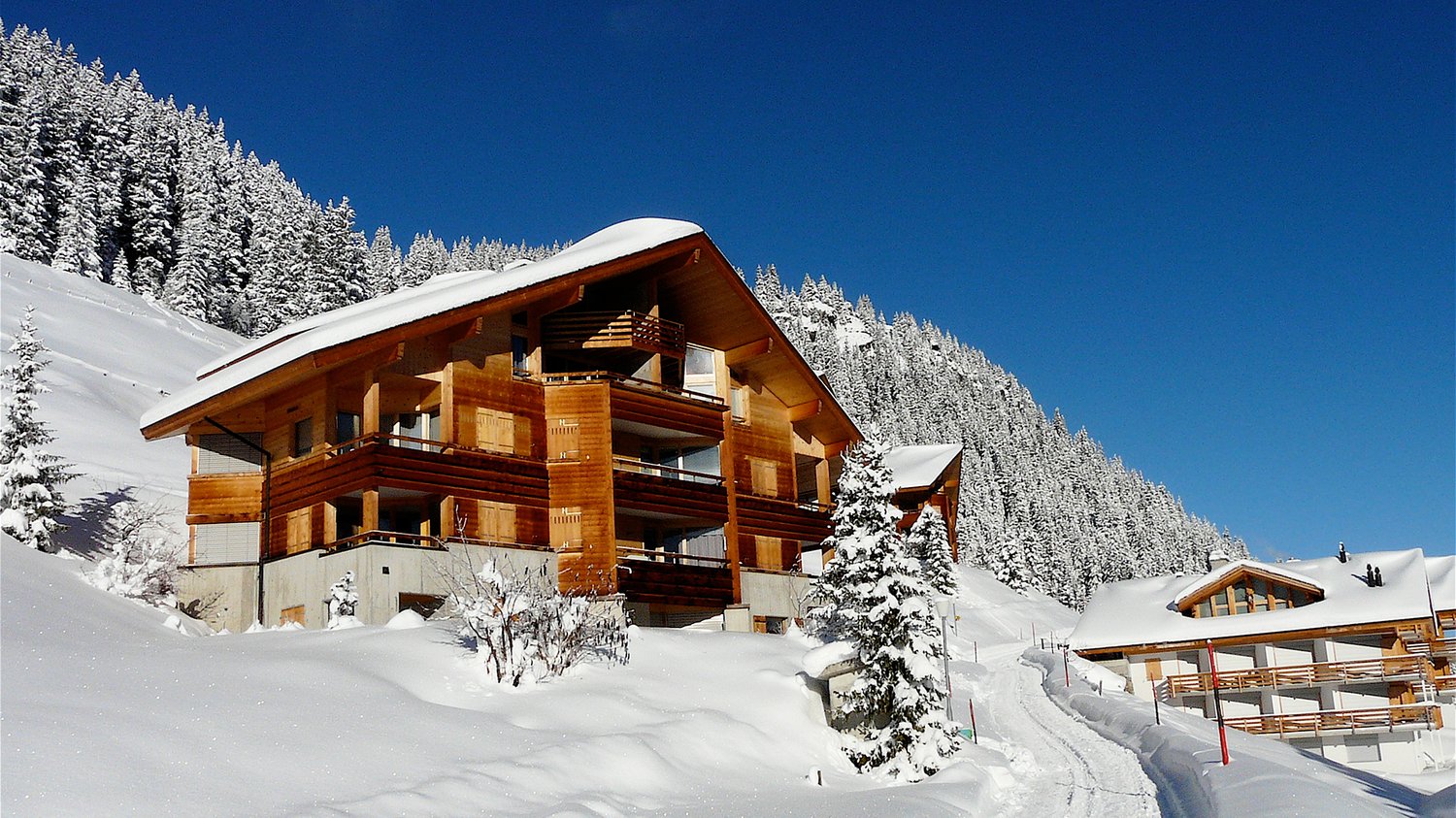 wooden chalet, multiple balconies, snow covered roof, surrounded by snow covered trees