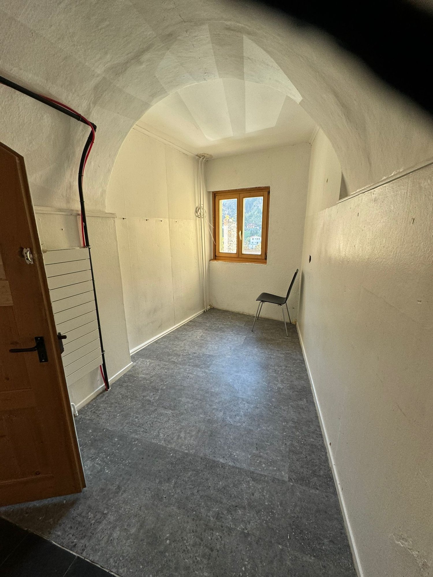 Empty room with arched ceiling, window, chair, wooden door, grey flooring, white walls, wires hanging on wall.