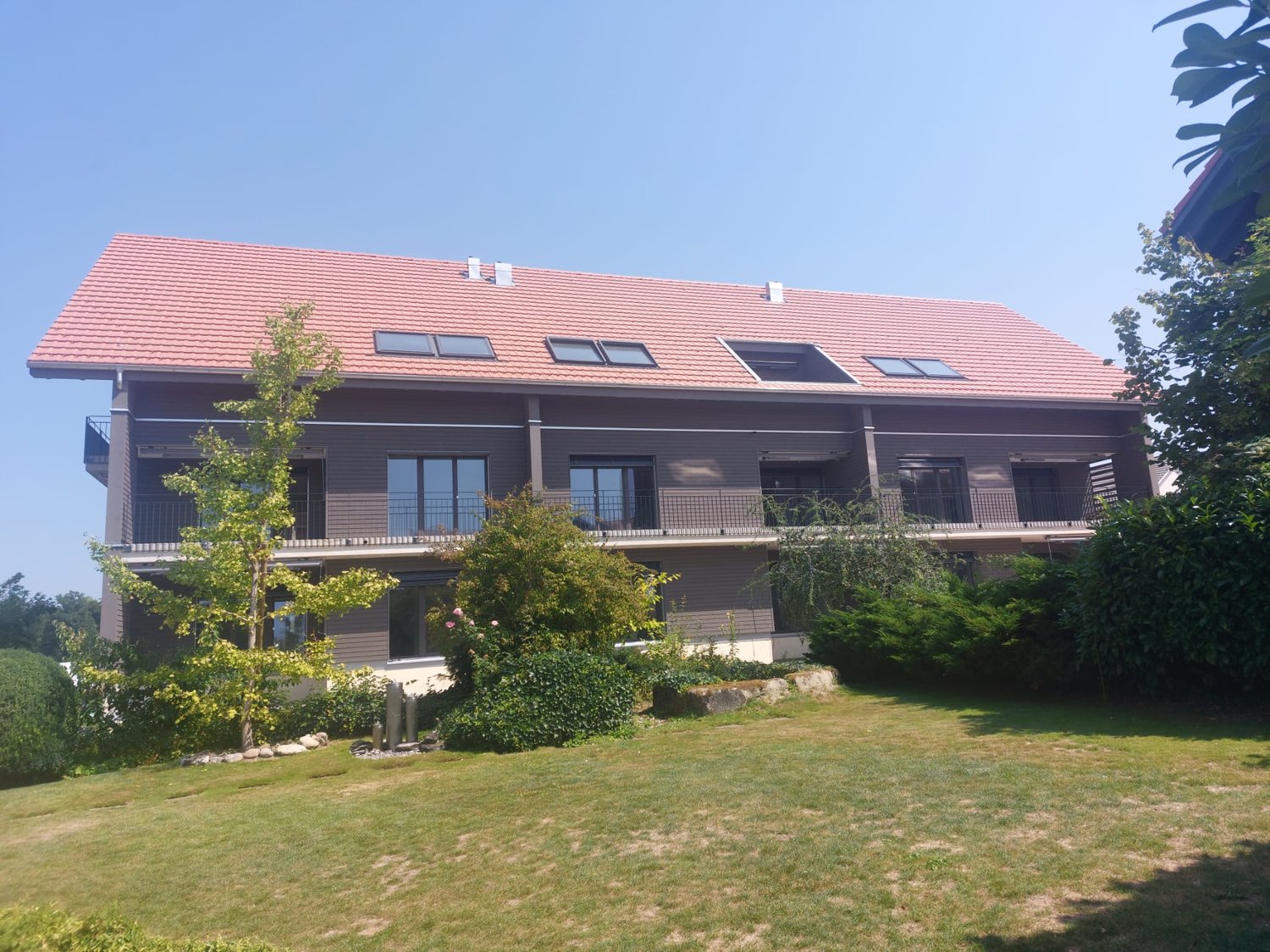 Two story house, red tile roof, brown walls, glass windows, balconies with railings, surrounded by plants and grass