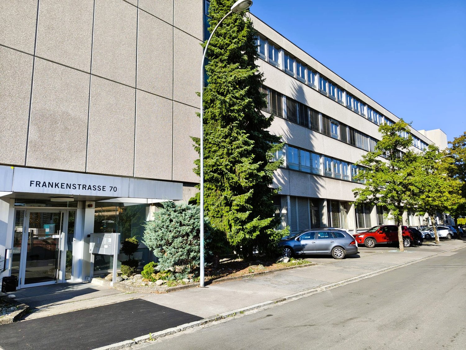 A modern building, likely an office building, with a glass door, name board reading 'Frankenstrasse 70', grey exterior, multiple windows, a few cars parked outside.