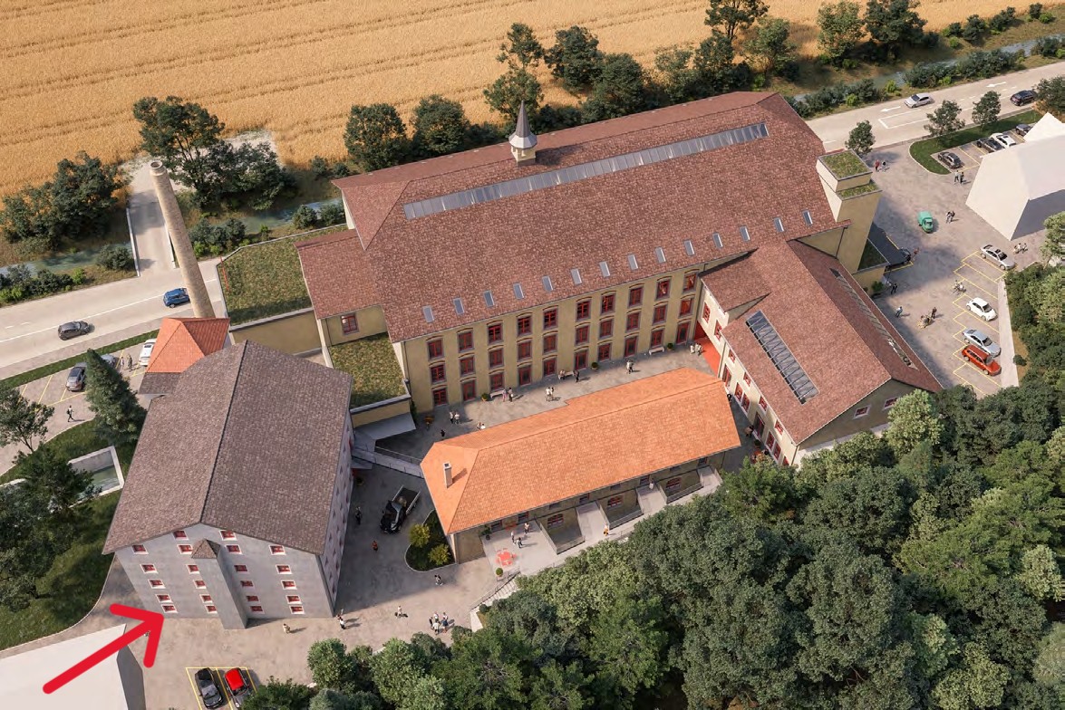 This image shows an aerial view of a large, multi-story brick building with a red tile roof. The building appears to be surrounded by trees and greenery, with a paved courtyard or plaza in front of it. There are several vehicles parked in the area around