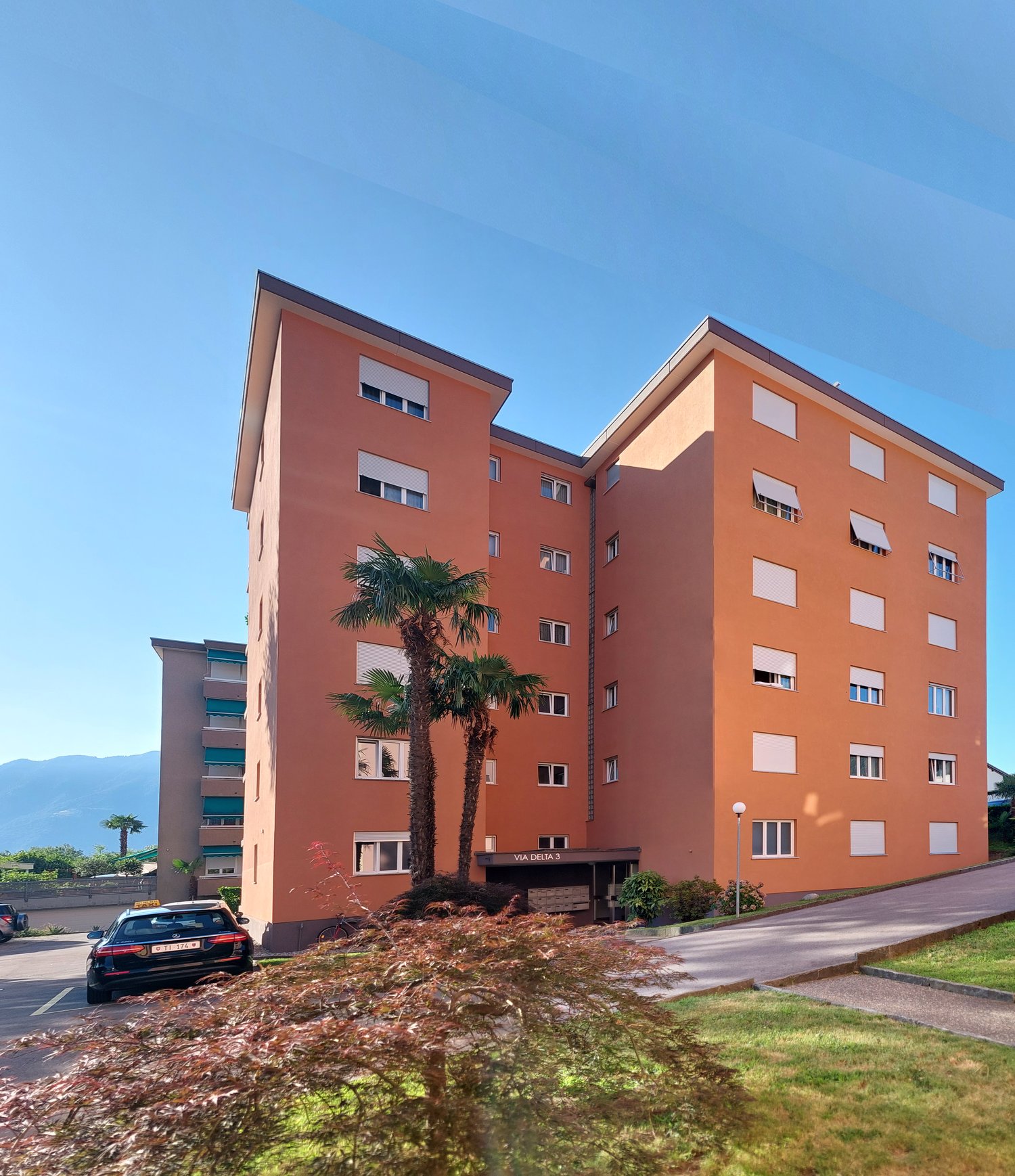 Multistory residential building, terracotta exterior, glass windows, two palm trees in front, parking lot with cars