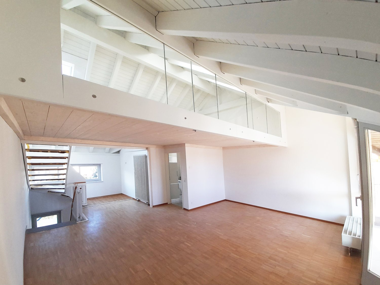 Empty room with hardwood floors, white walls, high wooden beams, glass railing, and an open staircase to the loft. Glass windows and an entrance door to the right.