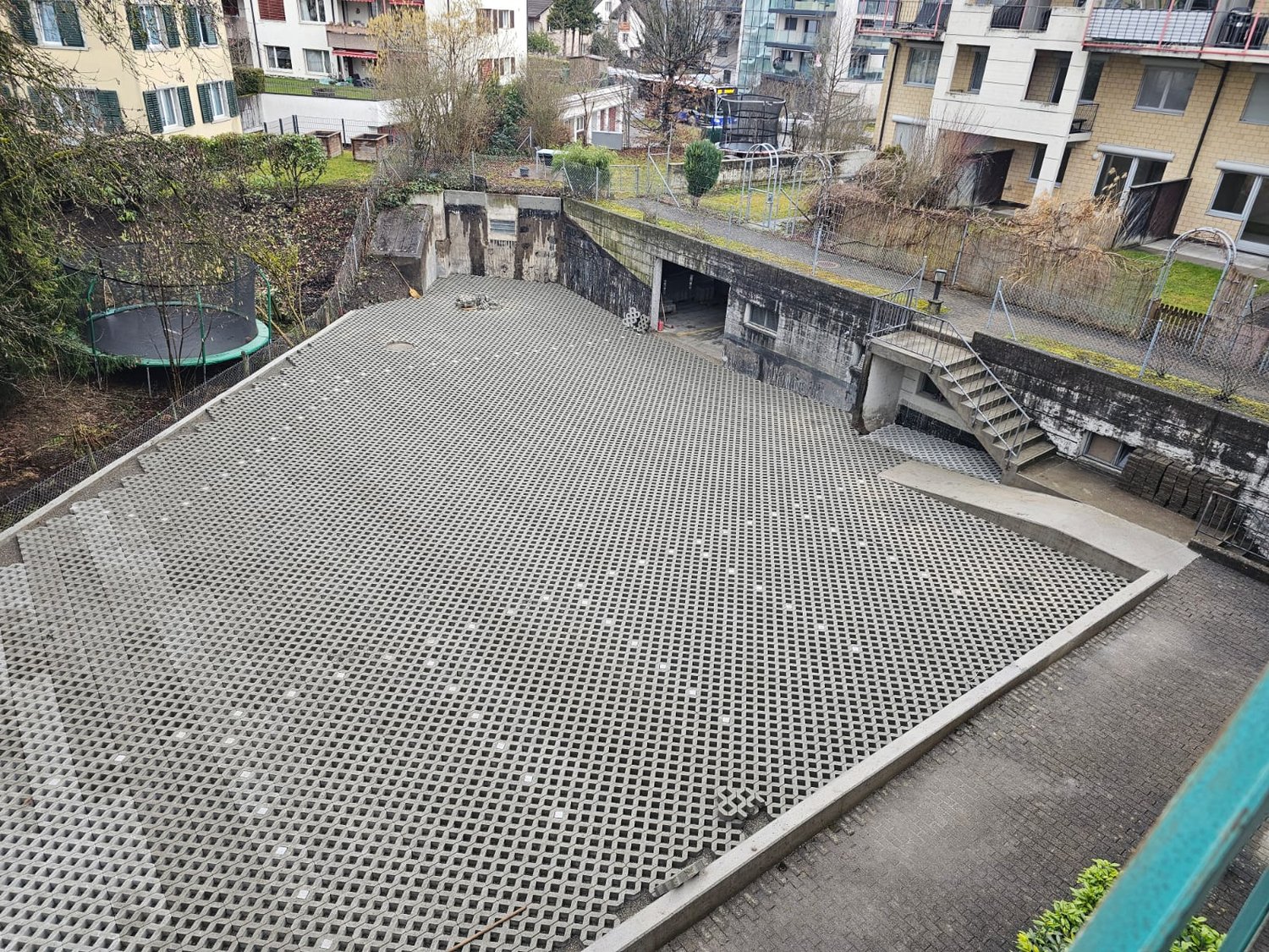 The image shows a raised concrete platform with a grid-like surface, likely serving as a walkway or outdoor area. There are stairs leading up to the platform, and the surrounding area appears to be a residential neighborhood with apartment buildings.