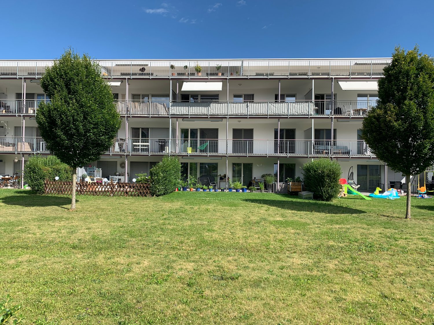 Modern apartment building with multiple floors and balconies, surrounded by a grassy area with plants and trees.
