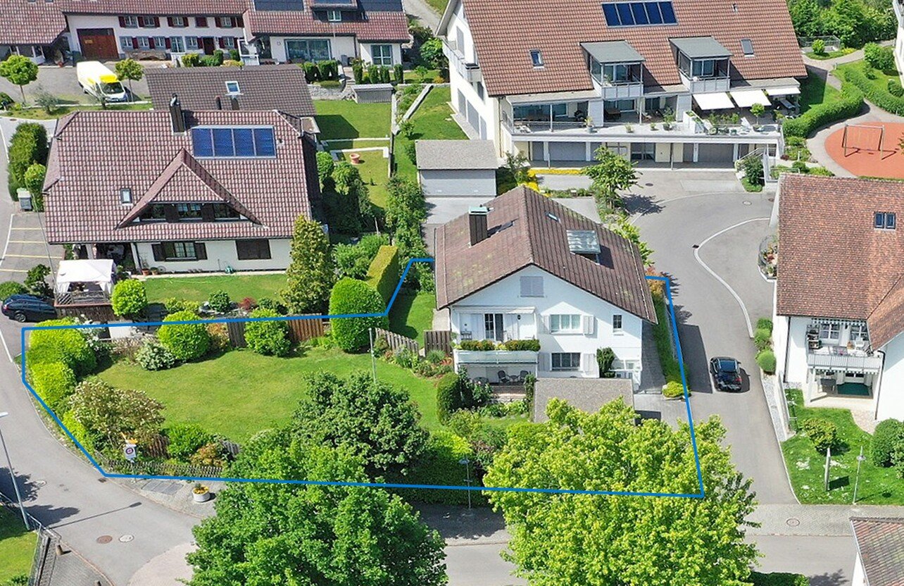 House with green roof, car parked in front, garden, solar panels, large grassy lawn, residential area, other houses and cars visible in the background