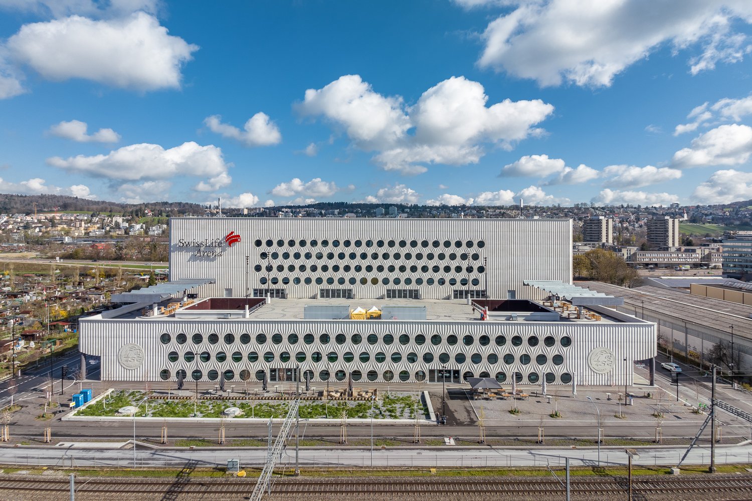 Large modern building with a distinctive white facade featuring a repeating pattern of circular openings. The building appears to be situated in an urban area with a view of the surrounding cityscape. There is a green landscaped area in the foreground of