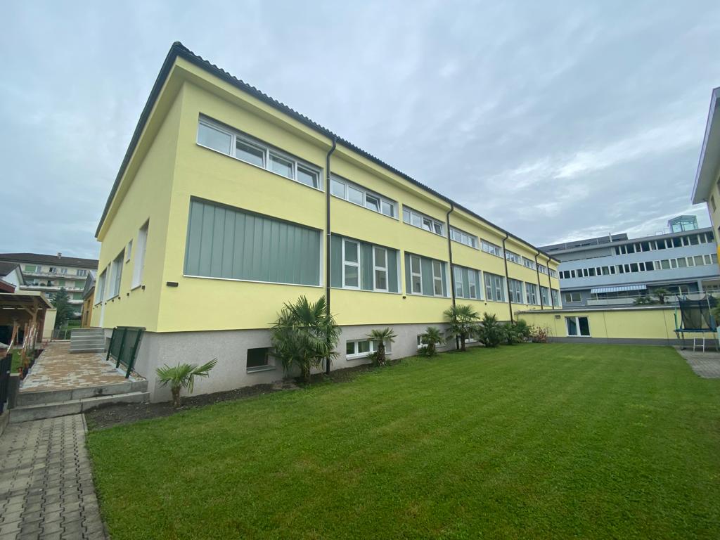 A yellow building with multiple glass windows and doors. There are several plants and bushes around the building, along with a wide green lawn. There is a concrete staircase in front of the building.