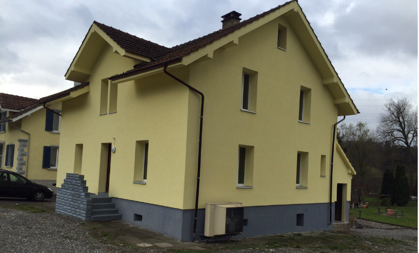 Two story house, yellow exterior, gray base, central chimney, steps leading up to the entrance, air conditioning unit