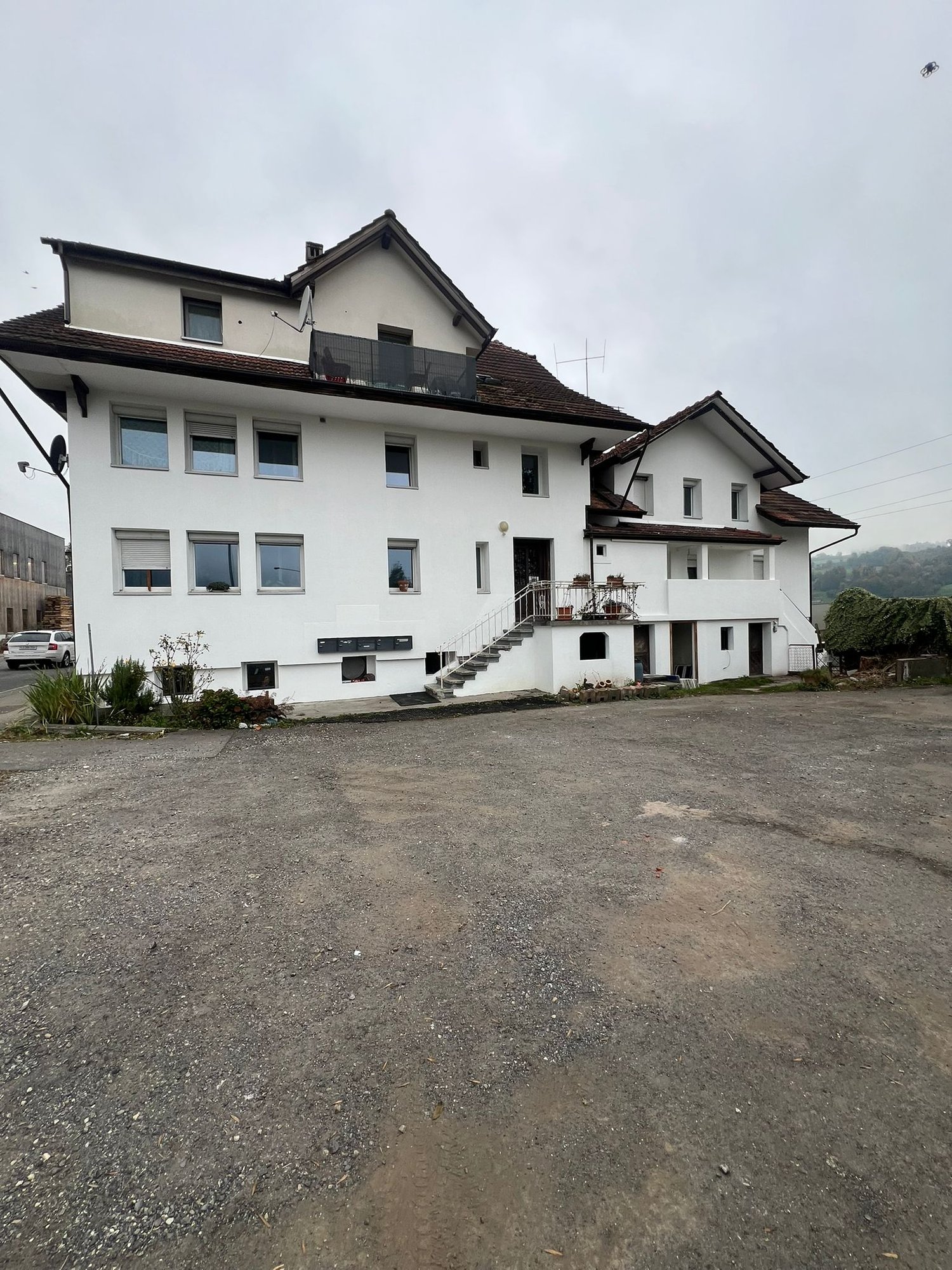 Two story house, white exterior, brown roof, multiple windows, balcony, staircases on both sides, car parked in front, large parking area in front, garden with plants and trees