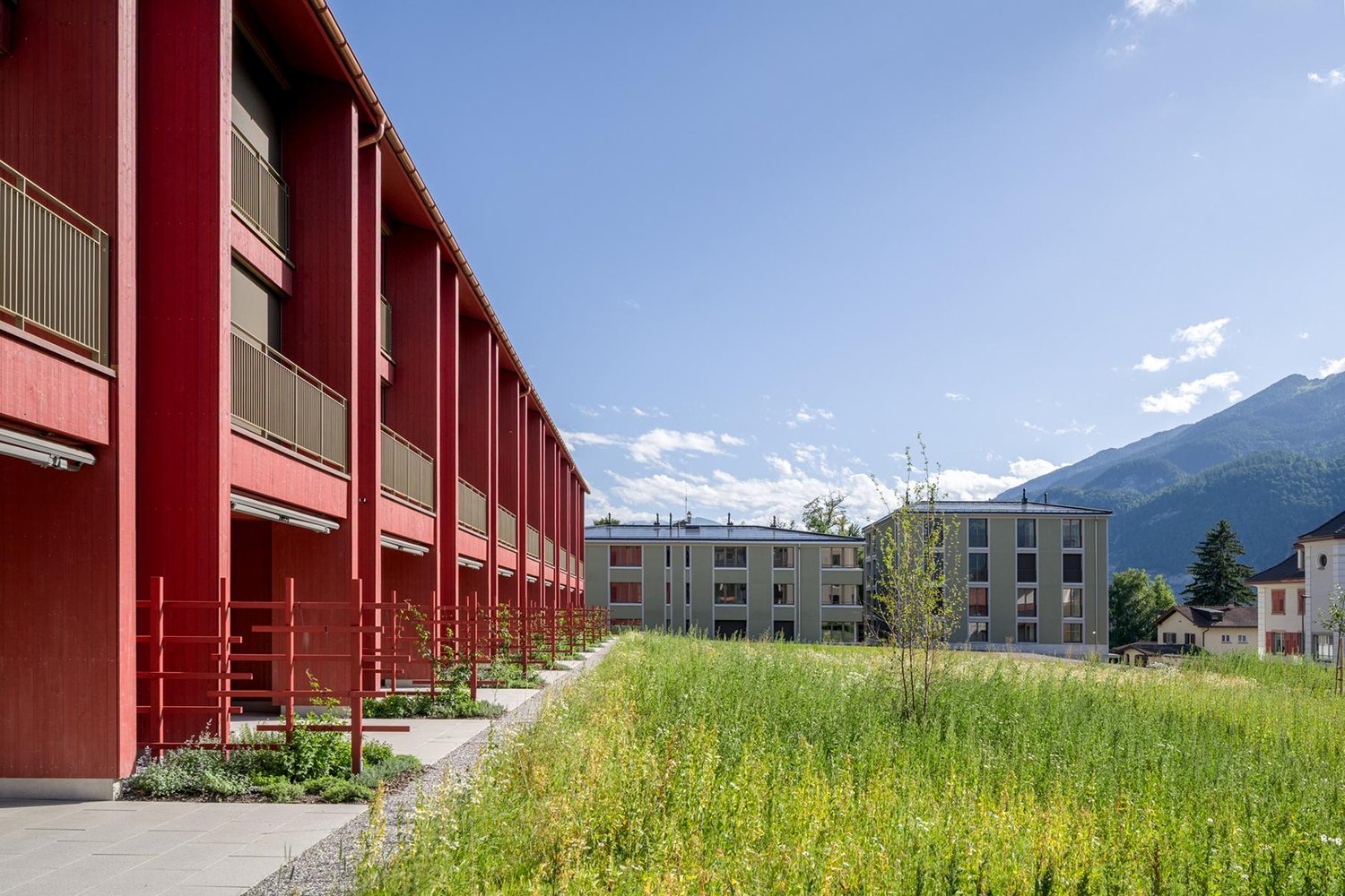 Multi-story apartment building with red exterior, balconies, surrounded by grassy area and trees, with mountains in the background