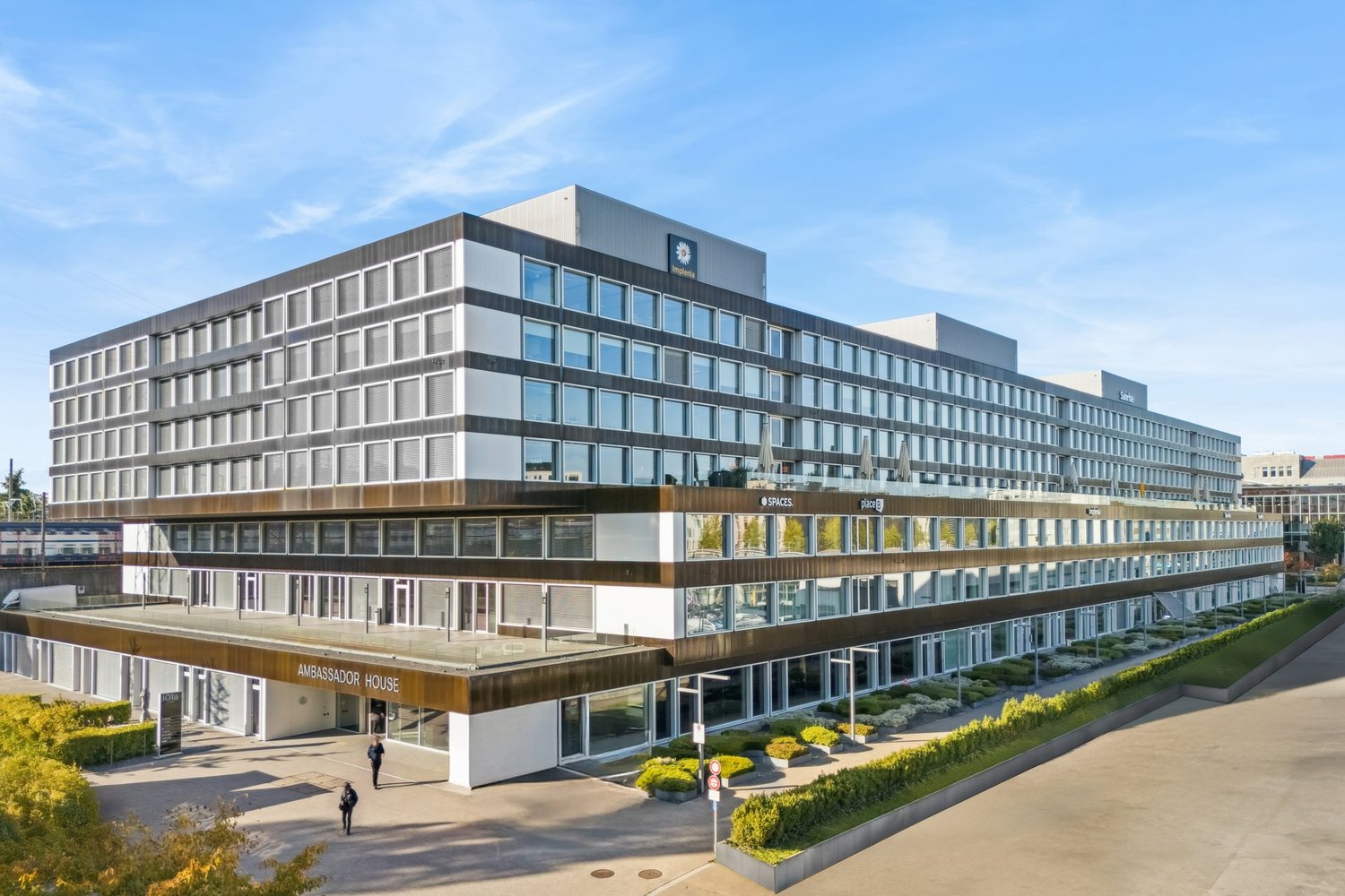 Modern office building with large glass windows, central entrance, balcony on the upper floor, multiple floors, and a green lawn in front.