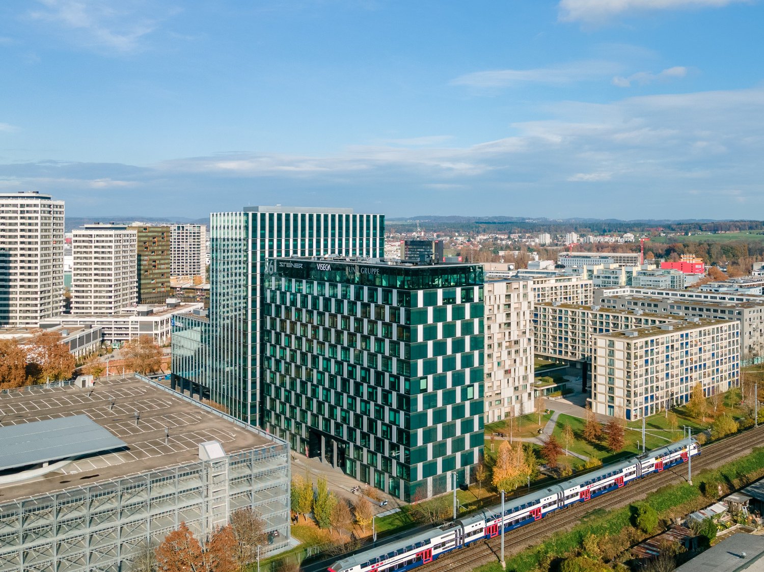 The image shows a cityscape with a mix of high-rise buildings, including a large green-and-white checkered office building in the center. There are also several other tall buildings, some with parking garages visible. A train can be seen running through t