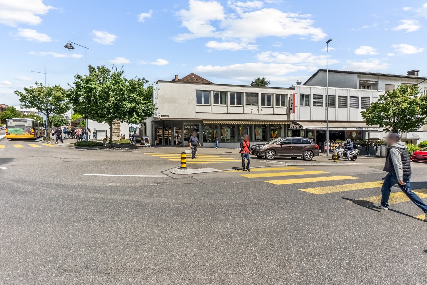 The image shows a busy street scene in an urban area. There are several buildings, including a cafe or shop, and people walking on the sidewalk. The street has yellow crosswalk markings and there are cars parked along the curb. The sky is partly cloudy.
