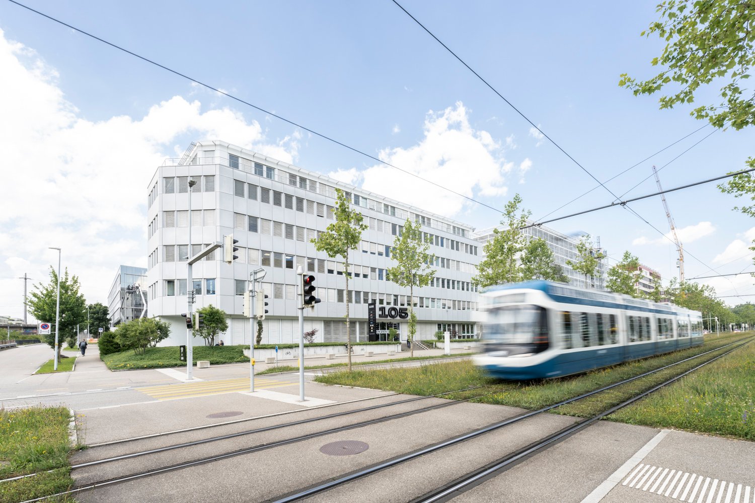 large building with many windows, tram tracks, green grass, trees, people, street lights, traffic light