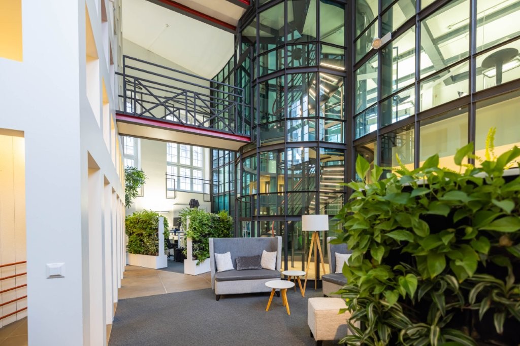 modern lobby, gray couch, white tables, large floor lamps, white wall, gray flooring, large plants, glass windows, staircase