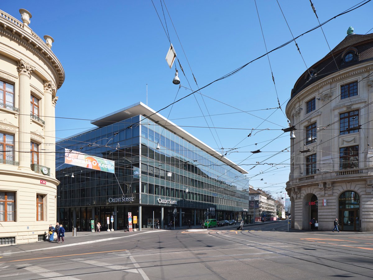 Large glass and steel commercial building with Credit Suisse branding, surrounded by historic ornate buildings, tram lines, and pedestrians on the street