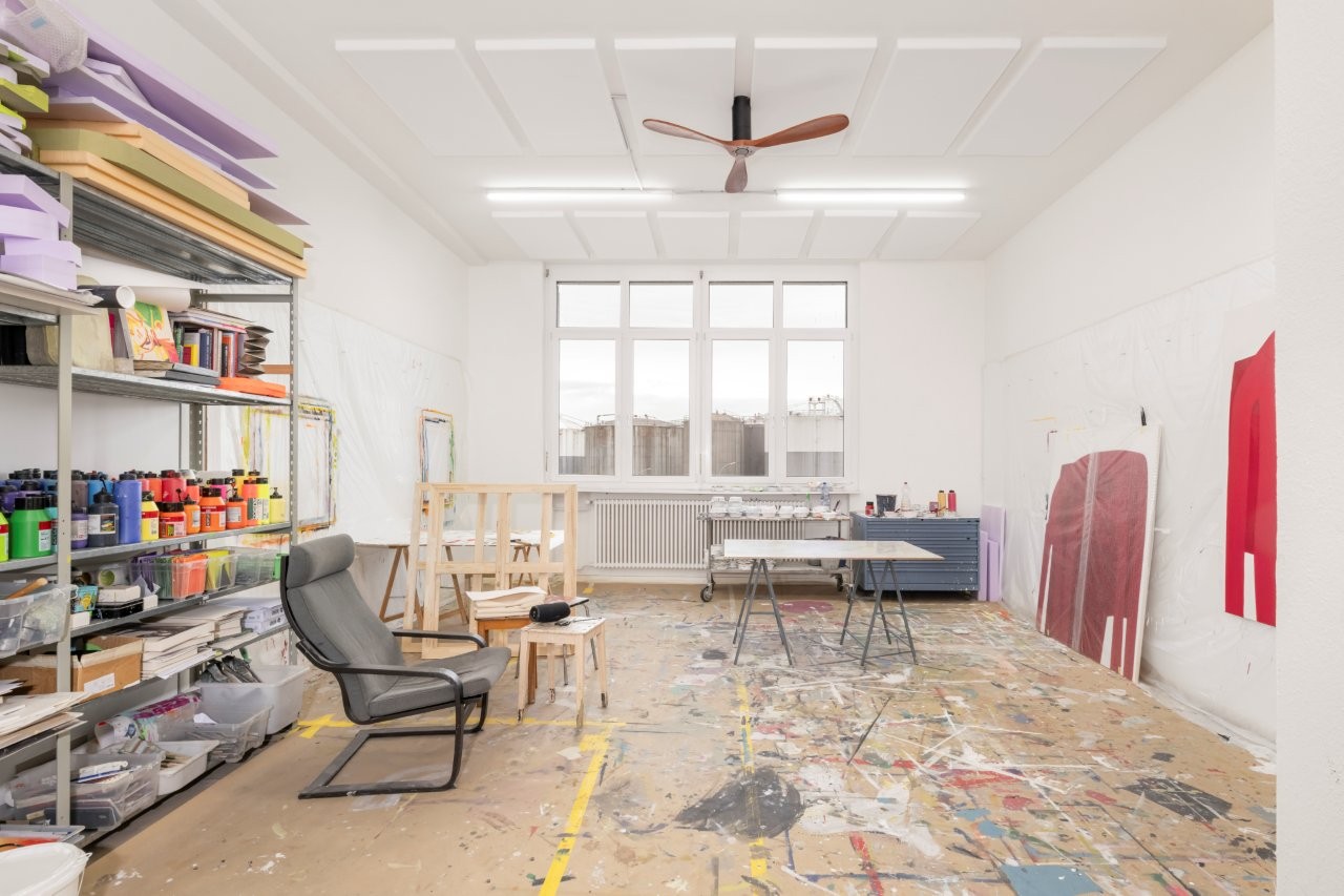 large room, white walls, ceiling fan, shelves with art supplies, tables, chairs, large windows, unfinished floor