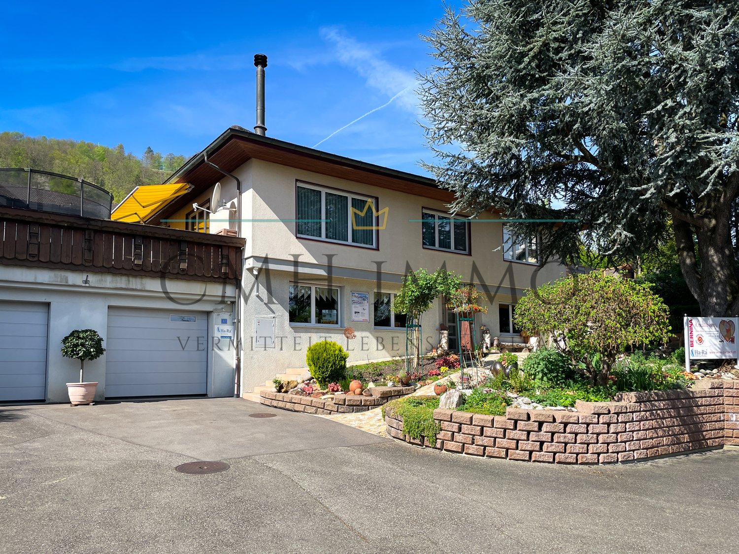2 story house with chimney, beige walls, gray garage, brick pathway, garden with trees and plants, large tree