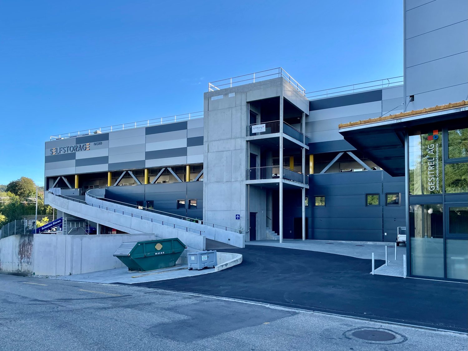 A multi-story commercial building with a gray and blue exterior. The building has a large garage door and a dumpster in the foreground, indicating it is likely a storage or warehouse facility.