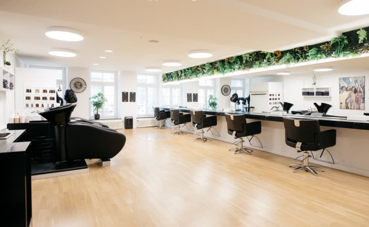 Hair salon with wooden floors, large windows, two black lounge chairs, a black workstation, two black barber chairs, a green wall design, and a painting on the wall.