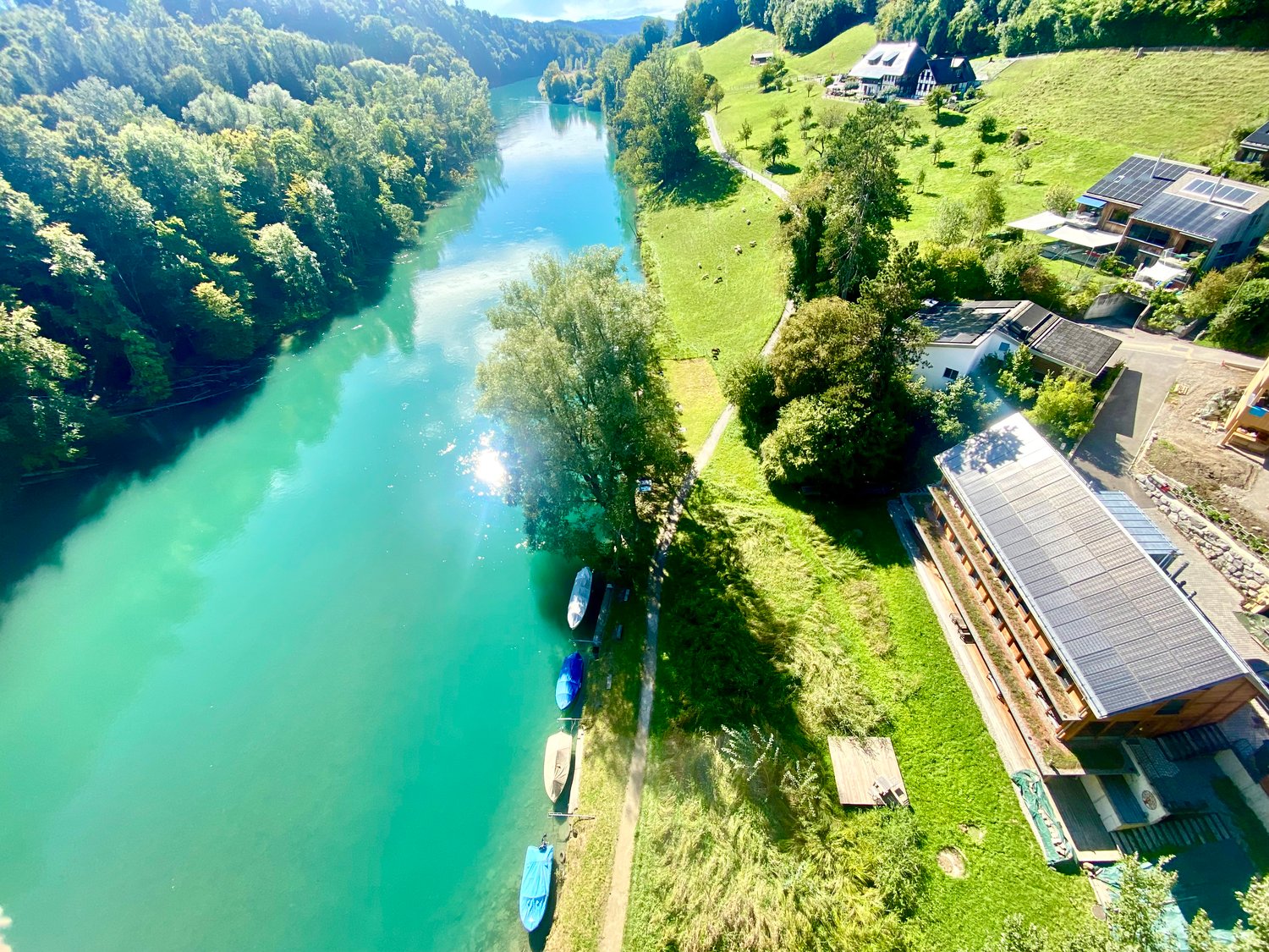 riverfront house with solar panels, multiple boats, grassy fields, mountains in the distance