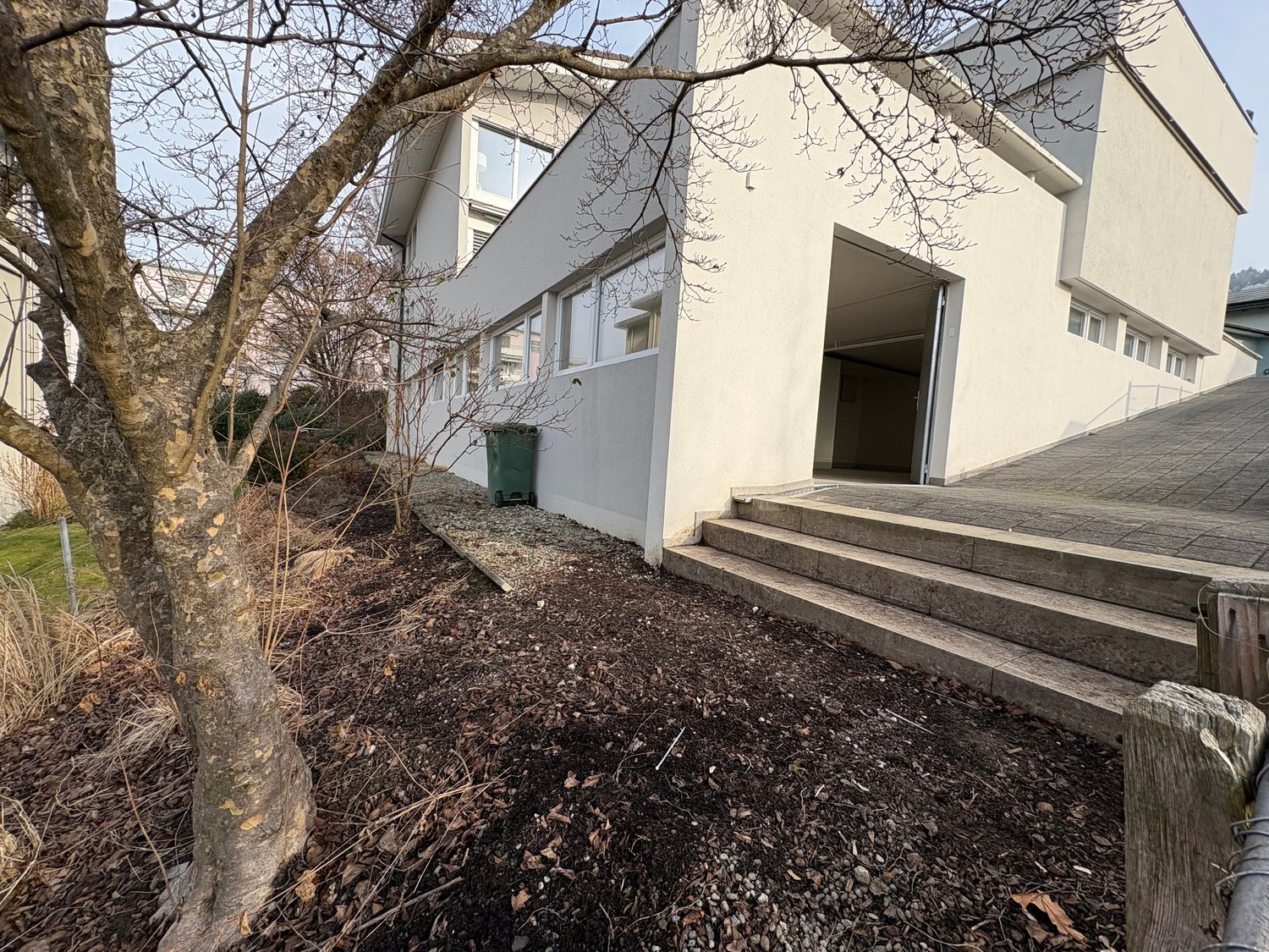 Modern architectural style, white facade, large windows, elevated ground floor, steps to the entrance, green container